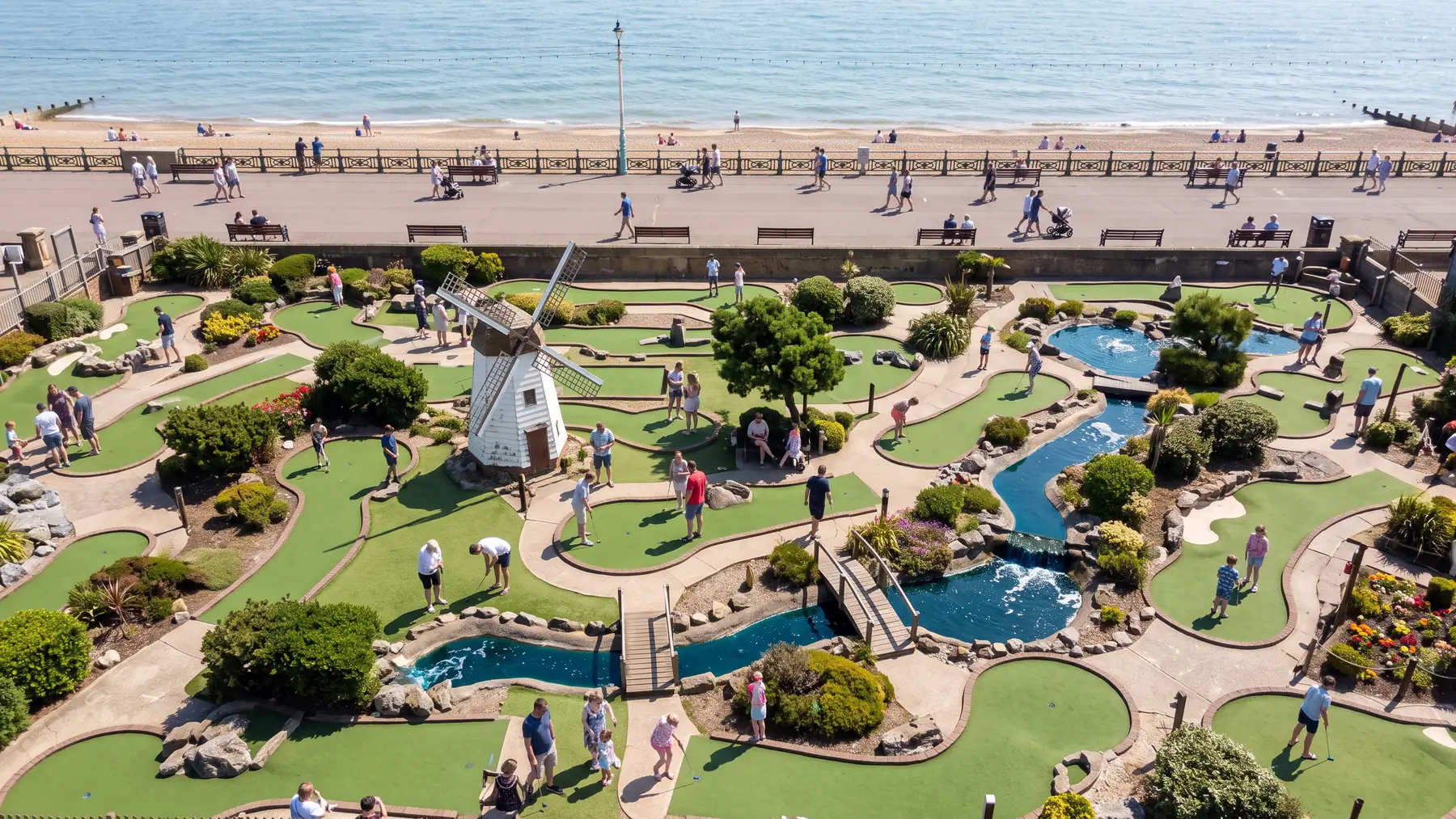 A busy seaside adventure golf course from above, with a windmill, water features and footbridges, the promenade and the sea beyond