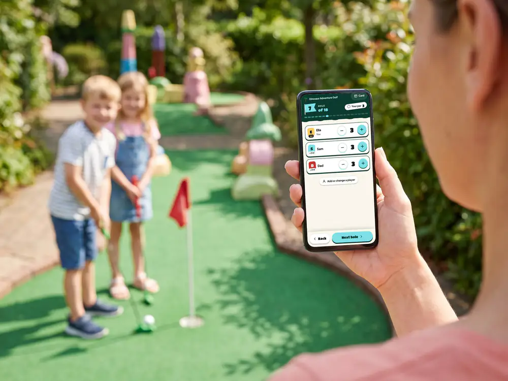 A phone held up on the course showing hole 7 of the scorecard, with a minus and a plus beside each player's score, as two children putt behind it