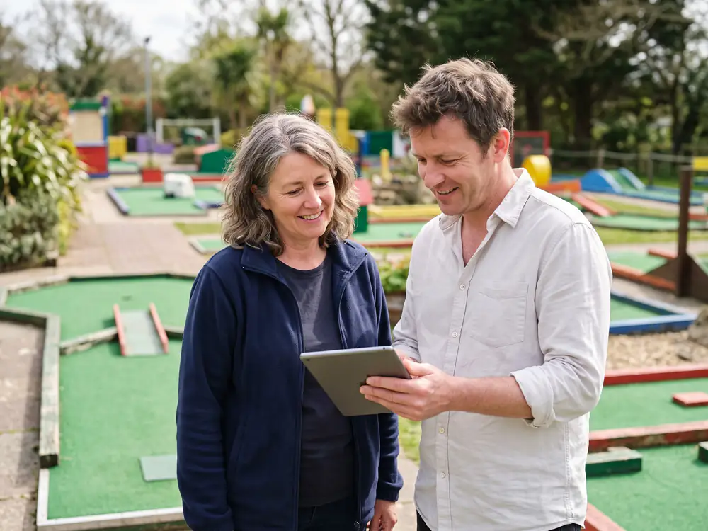 A course manager in a navy fleece looks at a tablet with a visitor on the course, both smiling