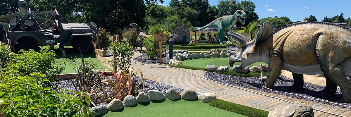 The marker for hole 17, a timber walkway between greens, an army truck with a dinosaur on the back, and a triceratops, a tyrannosaurus and a stegosaurus around them